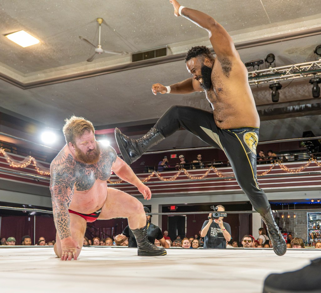 A professional wrestling match featuring a muscular athlete executing a kick on another wrestler who is on one knee. The scene is set in an indoor arena with an audience watching closely.