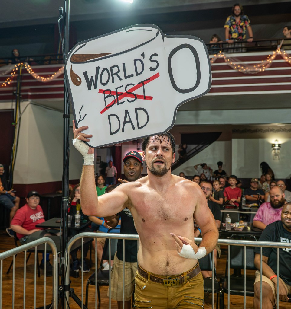 A shirtless male wrestler holding a large sign that humorously alters the phrase 'World's Best Dad' with a coffee cup illustration. He is pointing with a confident expression, surrounded by an audience in a wrestling venue.