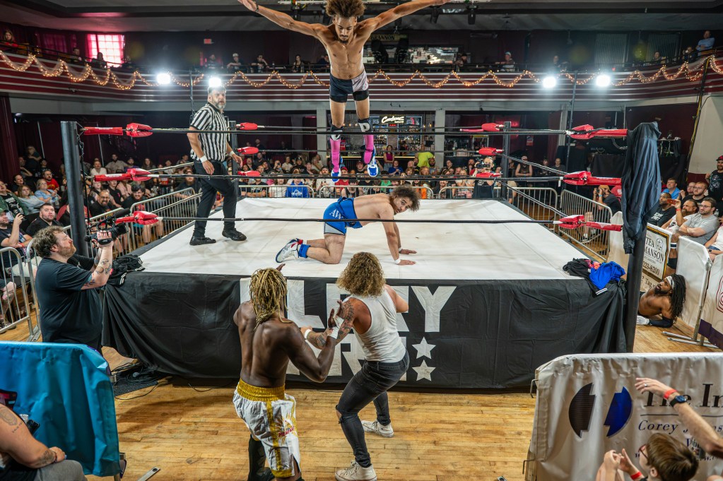 A wrestling event scene featuring a wrestler performing a leap from the ropes onto another wrestler in the ring, with a referee observing and a crowd in the background.