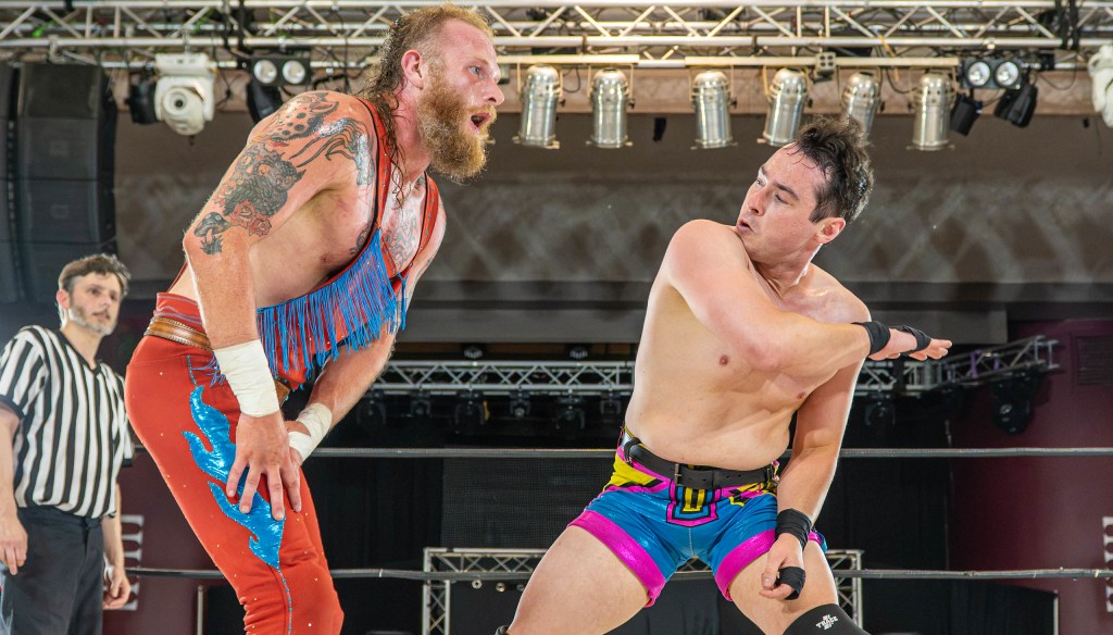 Two professional wrestlers in action, one with a beard and colorful attire, about to strike another wearing bright shorts, with a referee in the background.