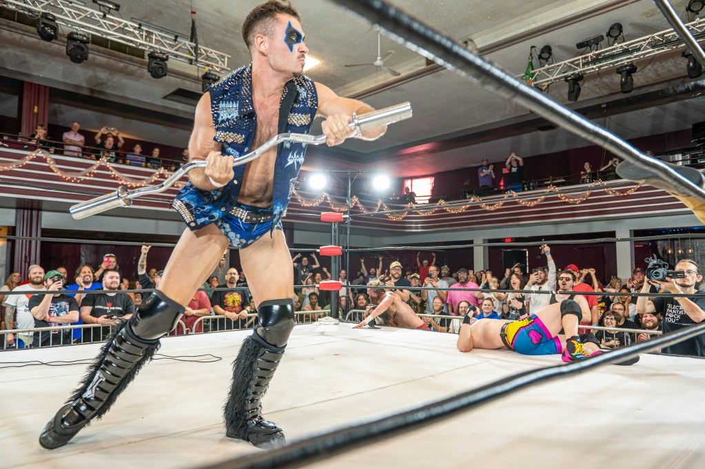 A wrestler in a flashy outfit holding a metal object stands in a wrestling ring, with an audience cheering in the background. Another wrestler is on the mat, while fans display excitement and engagement.