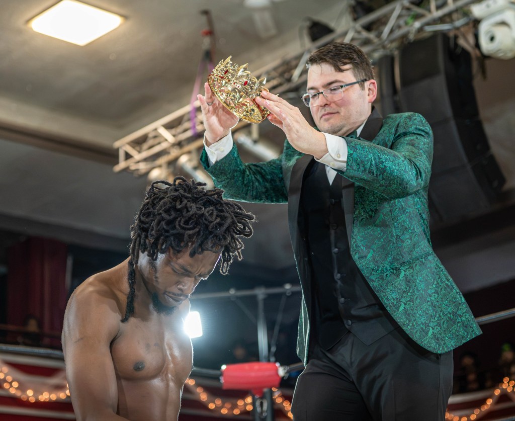 A man in a green patterned suit is placing a crown on the head of another man, who is shirtless with dreadlocks, in a wrestling ring.