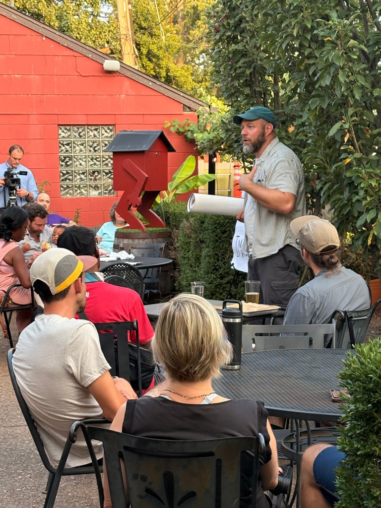A man with a beard in a green cap stands and gestures while speaking to an audience sitting at tables outdoors in a garden setting. A red wall and various plants are visible in the background.