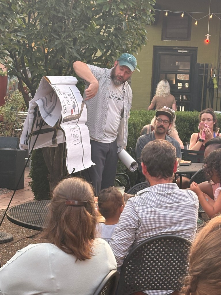 A man stands in front of an audience, holding a large scroll with illustrations, while a group of people, including children, watches attentively in an outdoor setting.