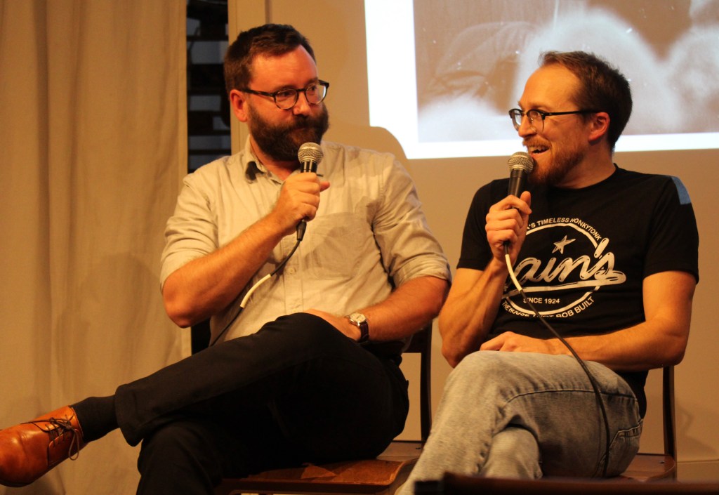 Two men seated on stage holding microphones, engaged in conversation during an event.