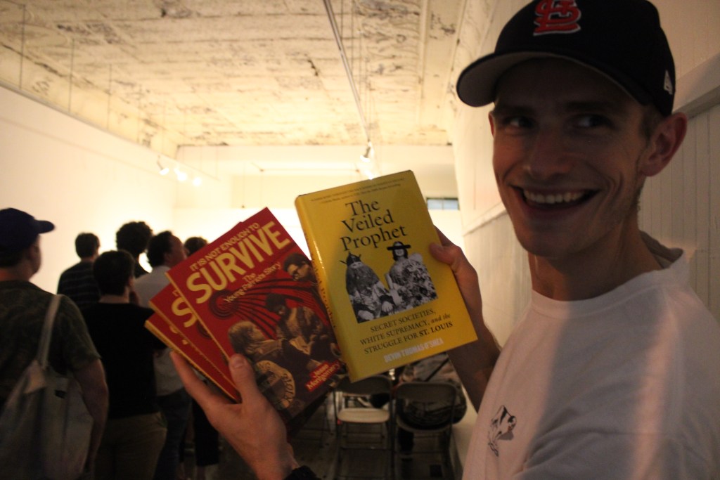 A man smiling while holding three books: 'It's Not Enough to Survive', 'Survive: Yours The Story', and 'The Veiled Prophet'.