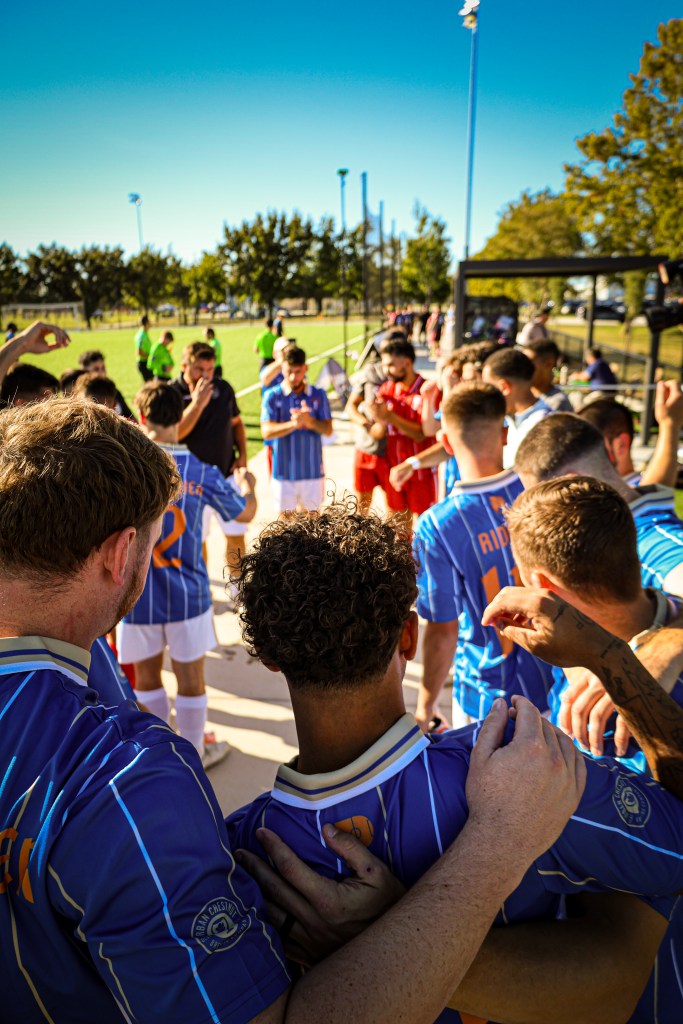 A group of soccer players in blue and red uniforms huddling together, celebrating or discussing strategy on a field with a clear blue sky in the background.