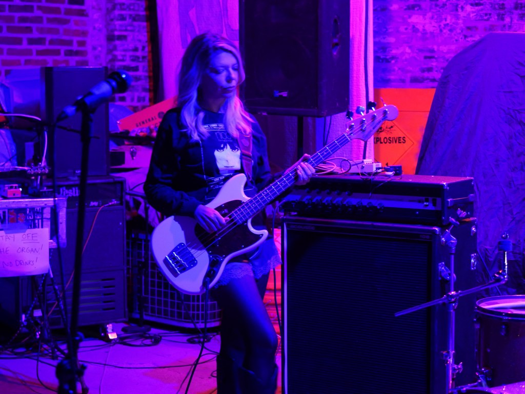 A bassist performing on stage with a microphone and amplifiers in a dimly lit venue exhibiting purple lighting.