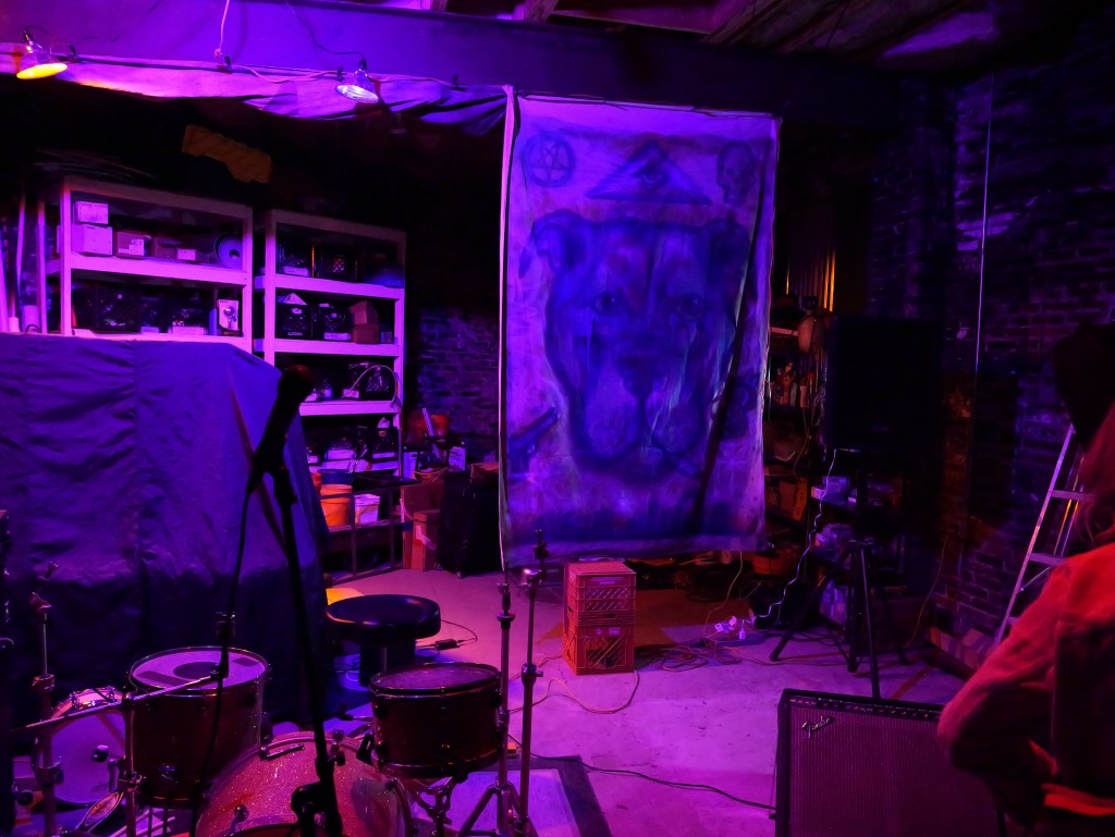 A dimly lit music venue with purple lighting, featuring a drum set, a microphone, and shelves filled with various items. A large banner with a dog illustration hangs prominently on the wall.