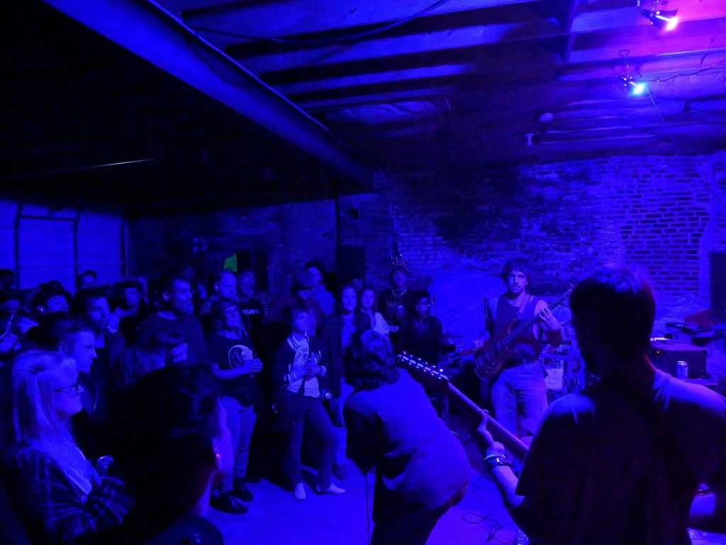 A crowd enjoying a live music performance in a dimly lit venue with blue lighting, featuring musicians on stage with instruments.