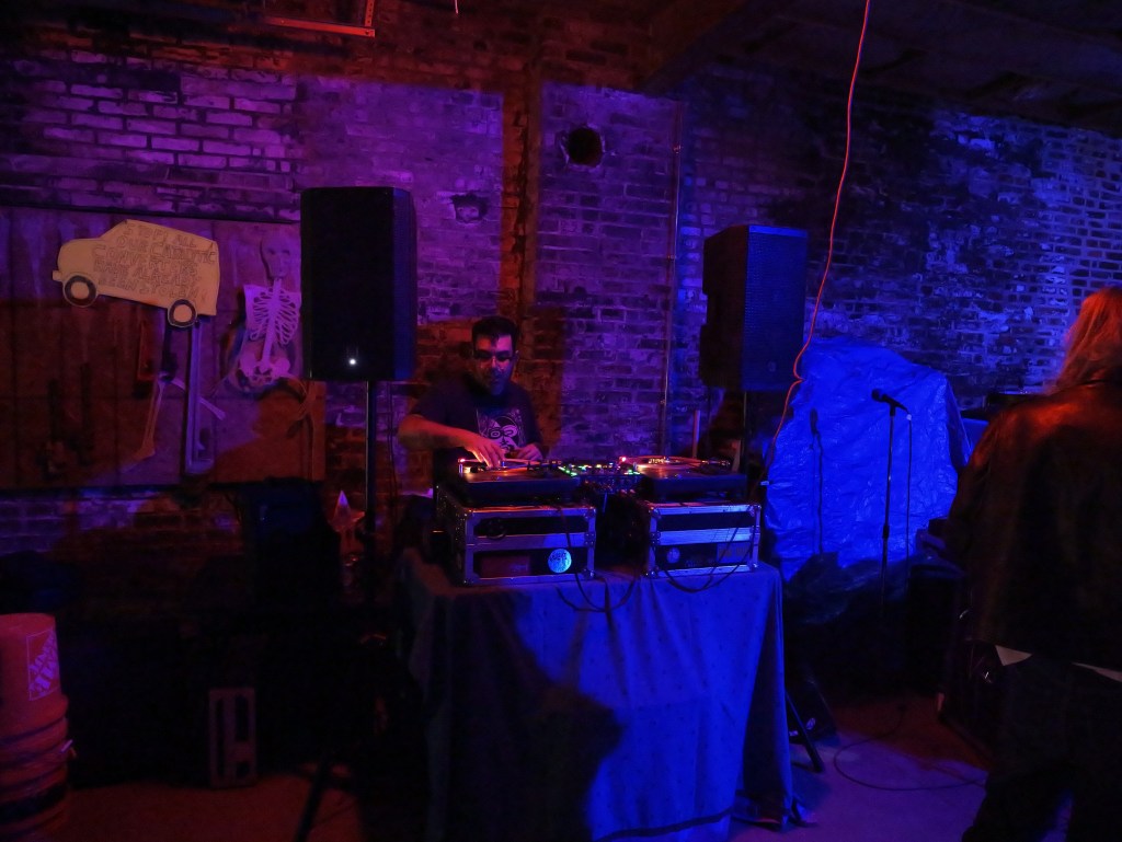 A DJ performing in a dimly lit venue with purple lighting, surrounded by speakers and music equipment, against a backdrop of exposed brick walls.