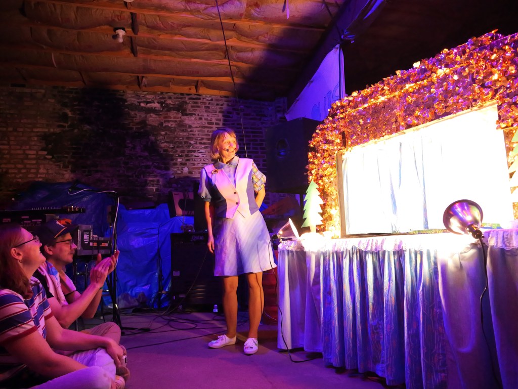 A performer in a light blue dress stands on stage in front of a decorated backdrop, interacting with an audience seated on the floor. The stage is illuminated with colorful lights, and a festive theme is present in the decor.