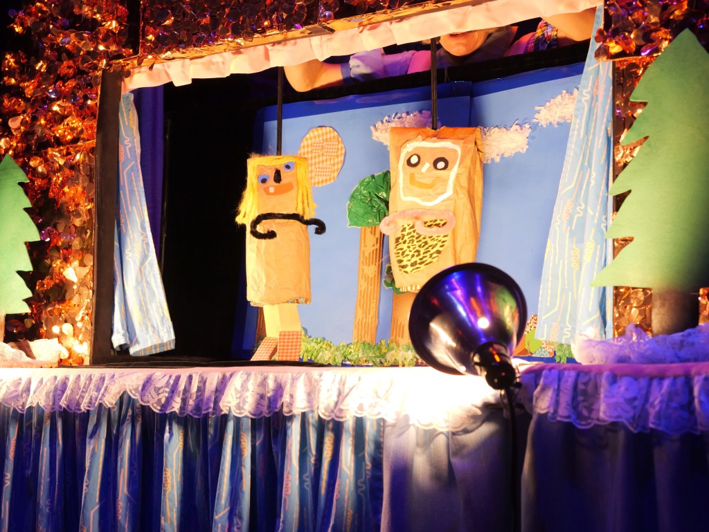 Colorful puppets featuring a girl with blonde hair and a character with a leopard print on a puppet theater stage with decorative lighting and a backdrop of trees and clouds.