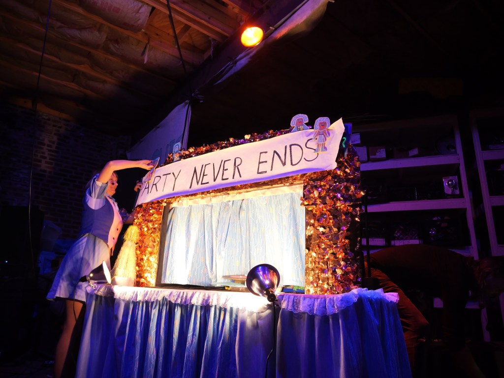 A performer adjusting a sign that reads 'PARTY NEVER ENDS' on a decorated stage with colorful lights and draped fabric.