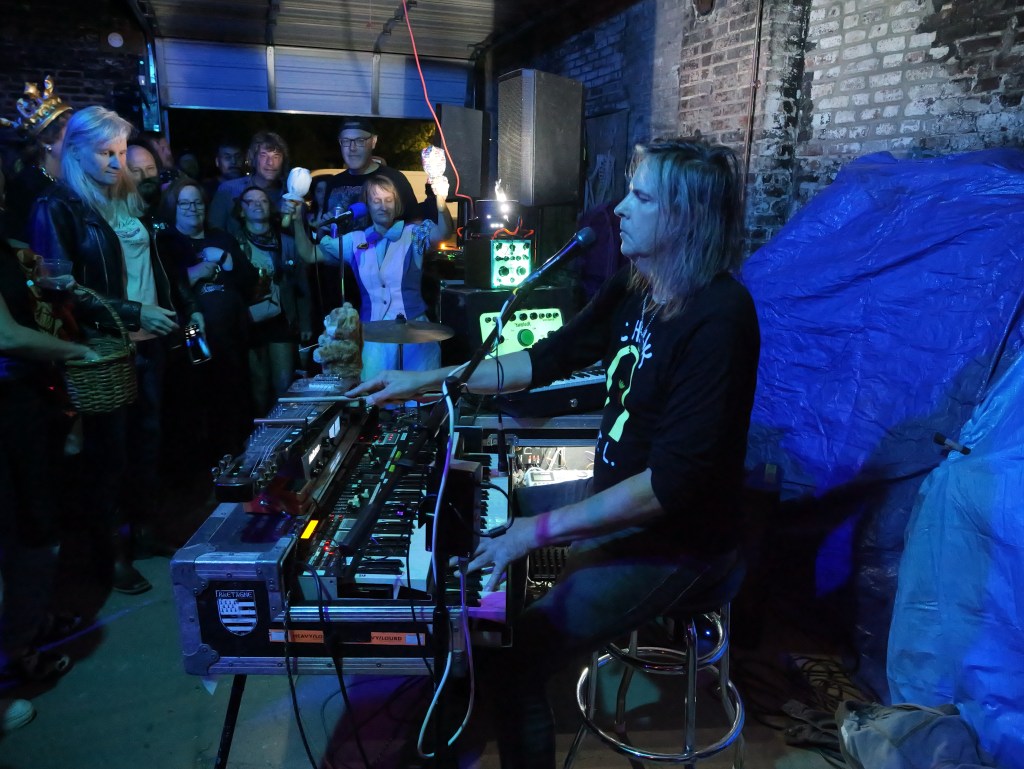 A musician performs at a small indoor venue, playing a keyboard setup while an audience watches attentively. The scene is illuminated with blue lights.