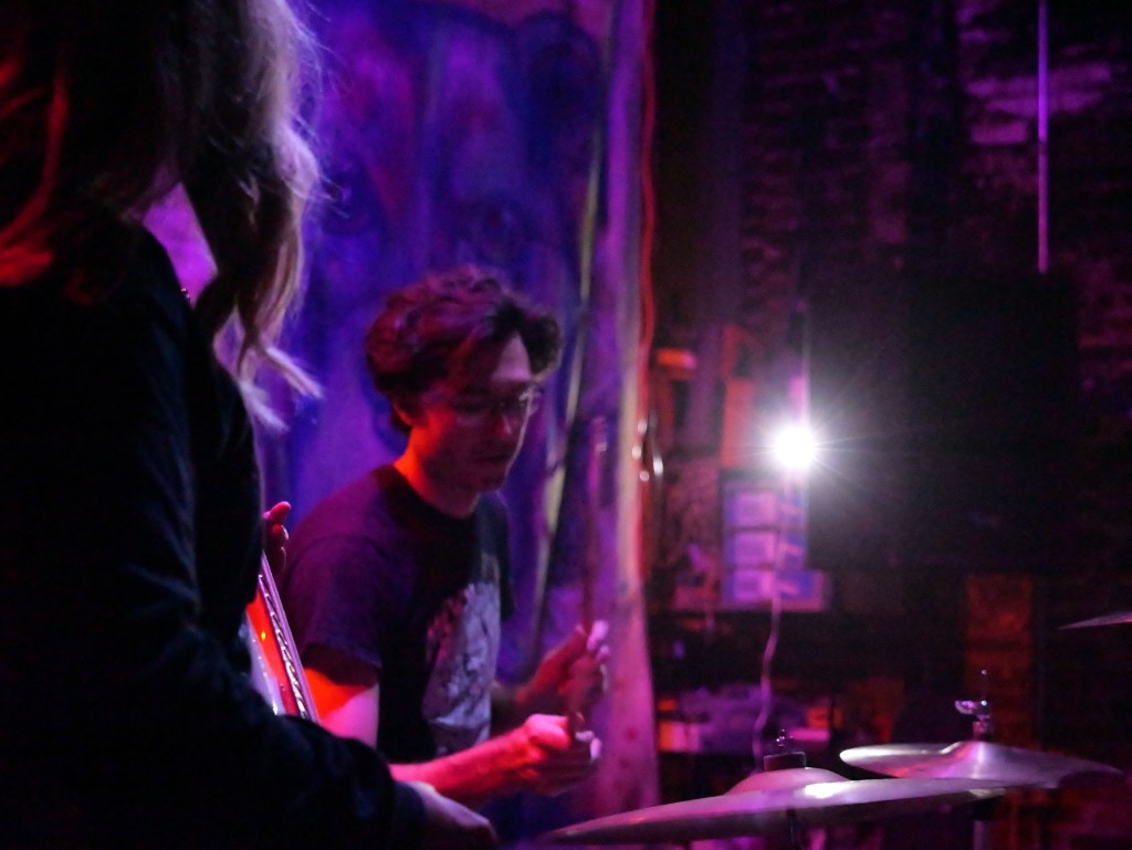 A musician playing the drums on stage, with soft purple and pink lighting, while another band member is partially visible in the foreground.