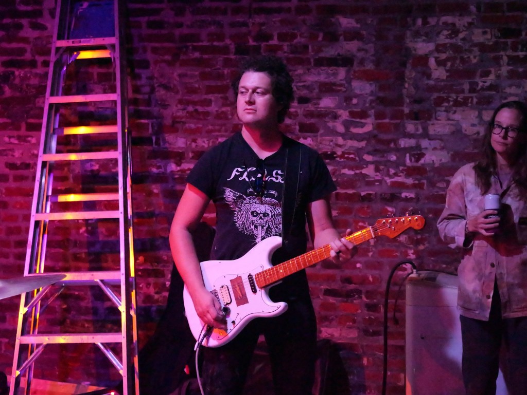 A musician playing an electric guitar on stage against a brick wall, with a ladder in the background and a woman in casual attire holding a drink.