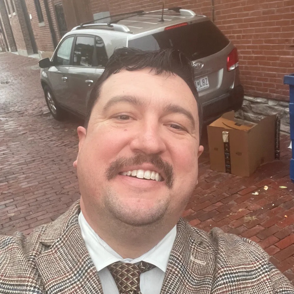 A smiling man taking a selfie outdoors, wearing a brown checkered suit with a white shirt and tie. In the background, there is a parked gray vehicle and a brick street.