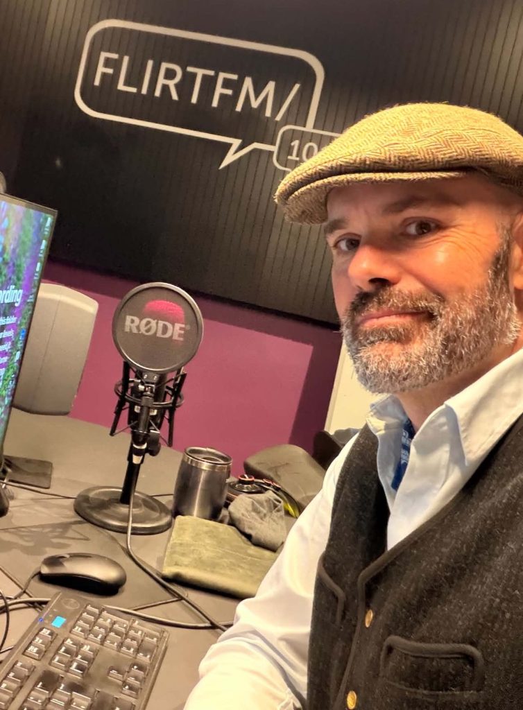 A man with a beard wearing a flat cap and a vest, sitting in a radio studio at FLIRTFM. A microphone, keyboard, and a computer screen are visible on the desk.
